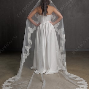 May include: A white wedding dress with a long, flowing skirt and a strapless bodice. The bride is wearing a long, sheer veil with intricate lace detailing along the edges. The veil is attached to a headpiece.