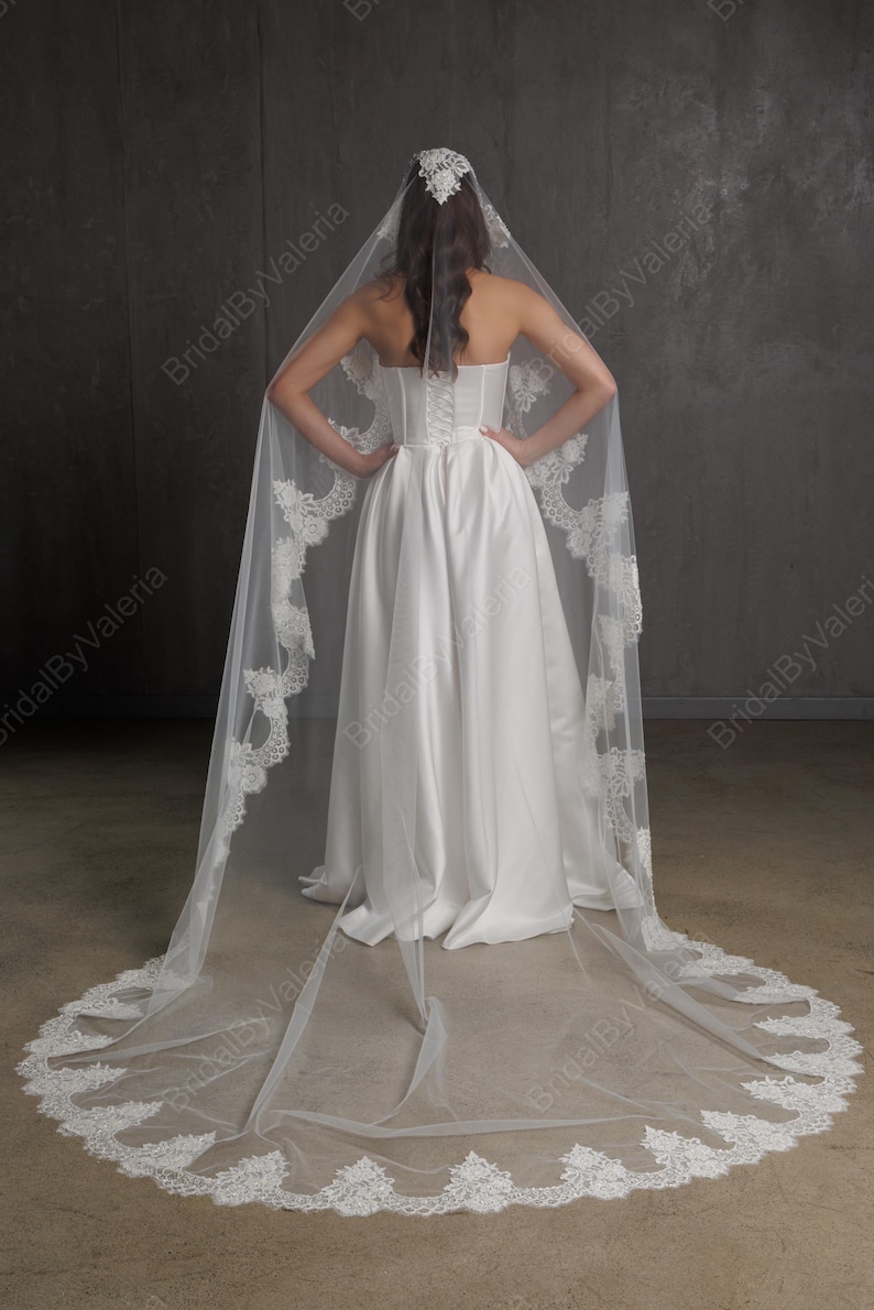 May include: A white wedding dress with a long, flowing skirt and a strapless bodice. The bride is wearing a long, sheer veil with intricate lace detailing along the edges. The veil is attached to a headpiece.