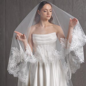 Puede incluir: Un velo de novia blanco con delicados bordes de encaje, drapeado sobre un vestido de novia blanco sin tirantes. El velo es transparente y el encaje a&ntilde;ade un toque de elegancia. El vestido tiene un corpi&ntilde;o ajustado y una falda fluida.