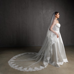 Puede incluir: Un vestido de novia blanco con una larga cola y un velo a juego. El velo presenta un delicado ribete de encaje en los bordes. El vestido tiene un corpi&ntilde;o sin tirantes y una falda fluida, creando una silueta elegante.