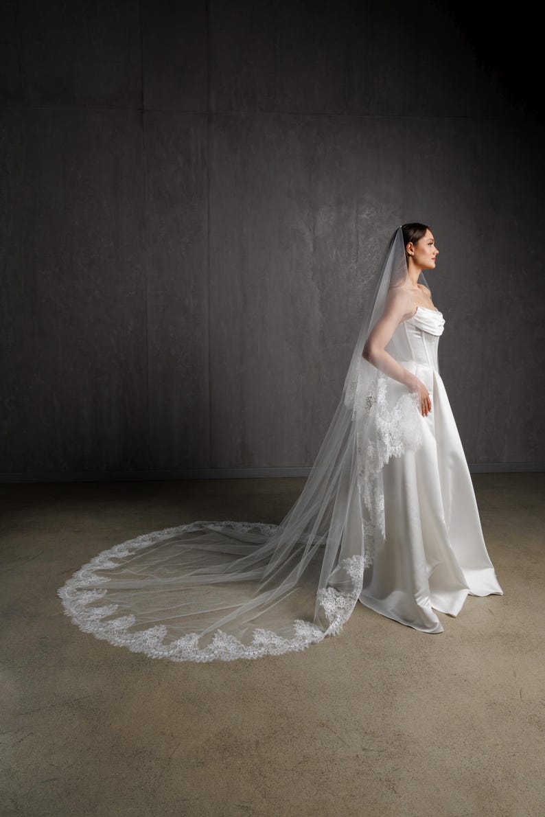 Puede incluir: Un vestido de novia blanco con una larga cola y un velo a juego. El velo presenta un delicado ribete de encaje en los bordes. El vestido tiene un corpi&ntilde;o sin tirantes y una falda fluida, creando una silueta elegante.