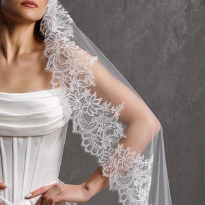 Könnte beinhalten: Ein weißer Brautschleier mit aufwendiger floraler Spitzenborte. Der Schleier ist über die Schulter drapiert und zeigt das filigrane Design. Die Braut trägt ein weißes Satin-Korsettkleid. Der Text "BridalByValeria" ist sichtbar.