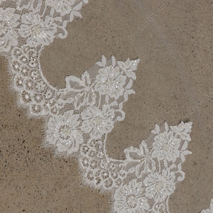 May include: Close-up of white floral lace with scalloped edges. The lace features detailed flower and leaf patterns, embellished with small, sparkling beads. The delicate design is set against a neutral, textured background, suitable for bridal or craft projects.
