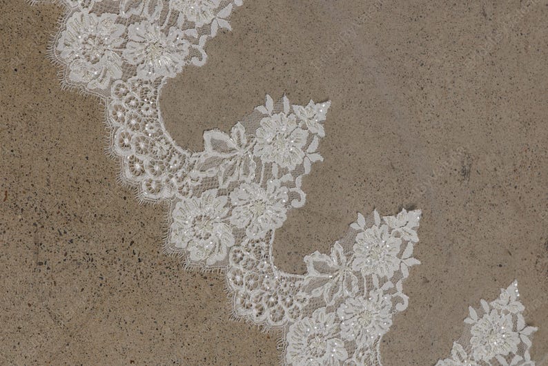 May include: Close-up of white floral lace with scalloped edges. The lace features detailed flower and leaf patterns, embellished with small, sparkling beads. The delicate design is set against a neutral, textured background, suitable for bridal or craft projects.