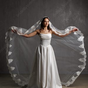 May include: A strapless white wedding dress with a corset bodice and a flowing skirt. The bride is wearing a long, sheer veil with delicate lace trim. The veil is spread wide, creating a dramatic effect.