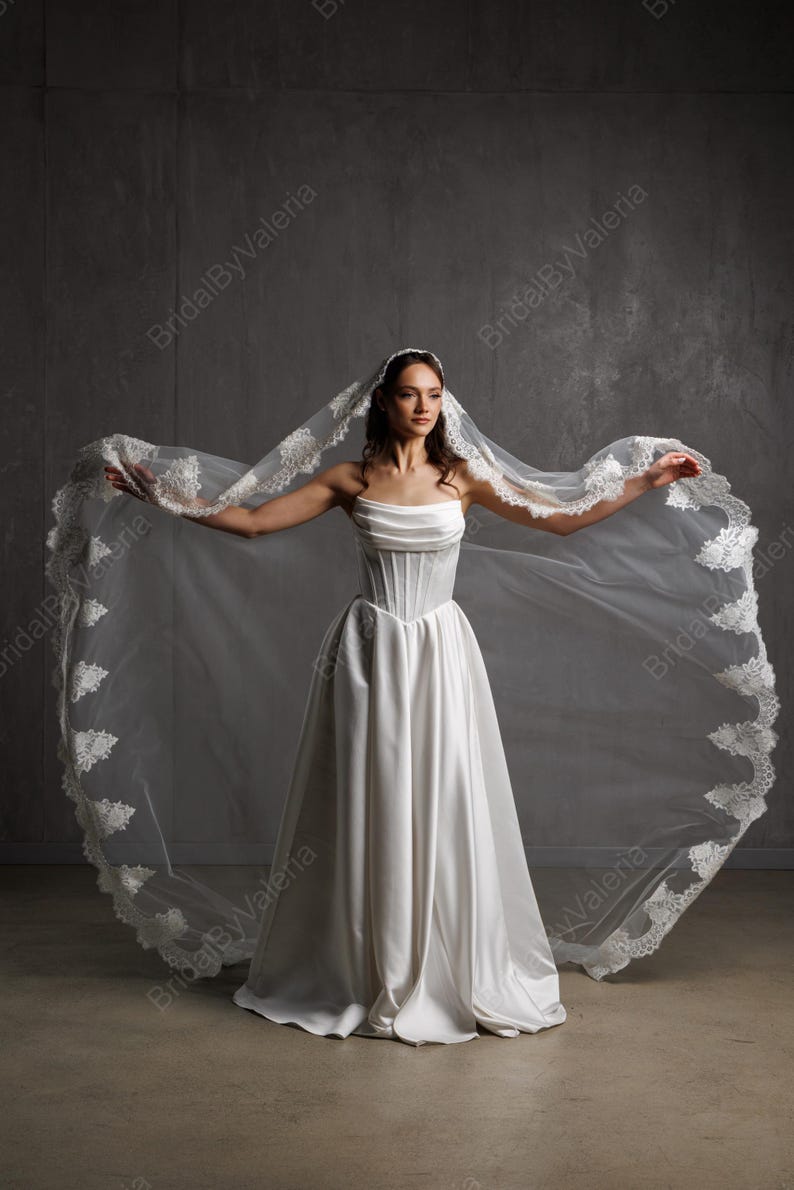May include: A strapless white wedding dress with a corset bodice and a flowing skirt. The bride is wearing a long, sheer veil with delicate lace trim. The veil is spread wide, creating a dramatic effect.