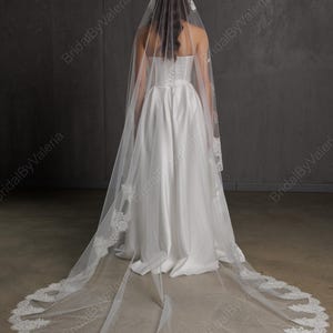 May include: A white bridal veil with a delicate lace trim and a matching lace headpiece. The veil is long and flowing, extending to the floor. The bride is wearing a white satin wedding dress. The background is a neutral gray.