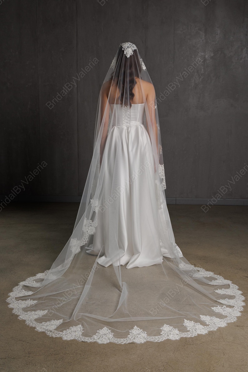 May include: A white bridal veil with a delicate lace trim and a matching lace headpiece. The veil is long and flowing, extending to the floor. The bride is wearing a white satin wedding dress. The background is a neutral gray.