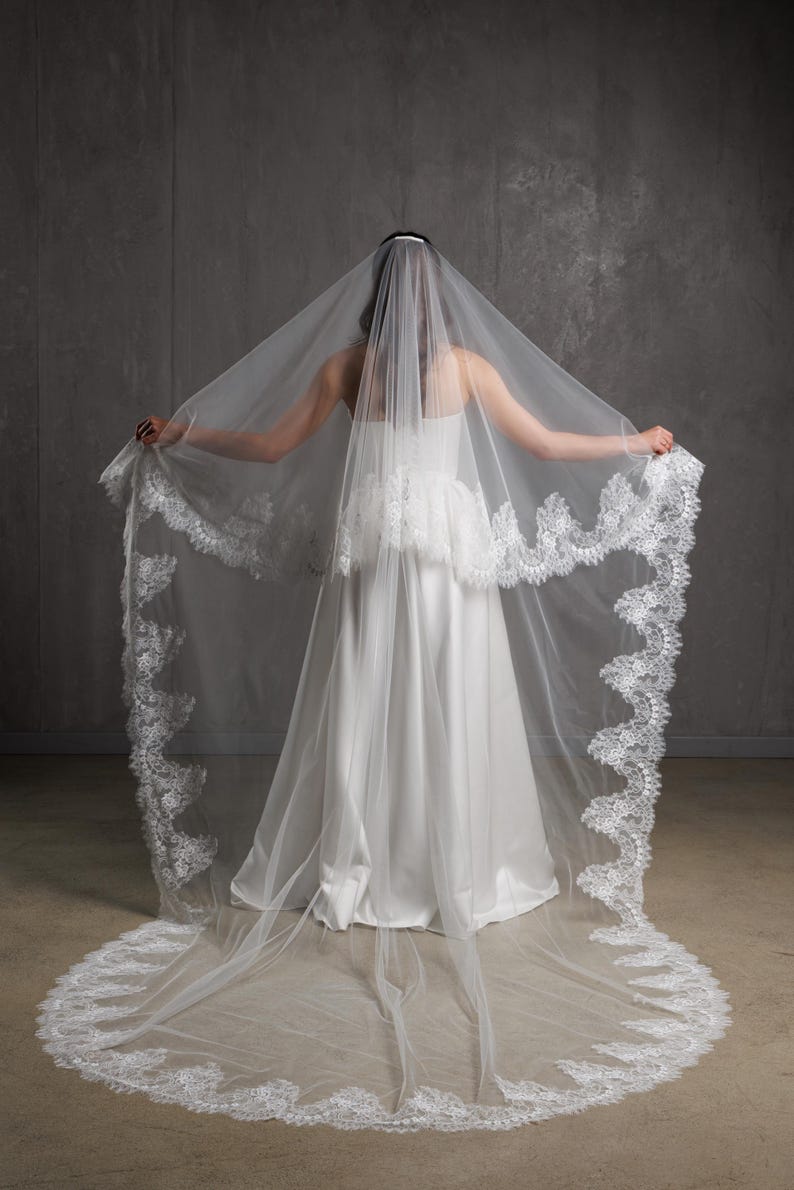 Puede incluir: Un velo de novia blanco con un delicado ribete de encaje. El velo se extiende, mostrando toda su longitud y dise&ntilde;o. La novia lleva un vestido de novia blanco, creando un look nupcial cl&aacute;sico.