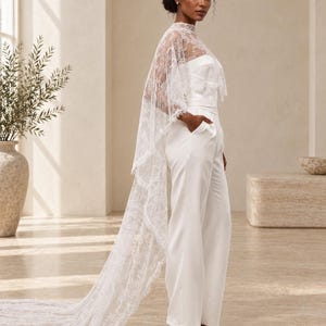 May include: A white strapless jumpsuit with wide legs and pockets, paired with a sheer, floor-length white lace cape featuring a high neckline and intricate floral patterns that drape down the back to form a long train on the floor.
