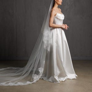 Puede incluir: Vestido de novia de sat&eacute;n blanco con corpi&ntilde;o sin tirantes y falda en l&iacute;nea A. Un velo largo y transparente con ribete de encaje cae por la espalda. El vestido es elegante y cl&aacute;sico.