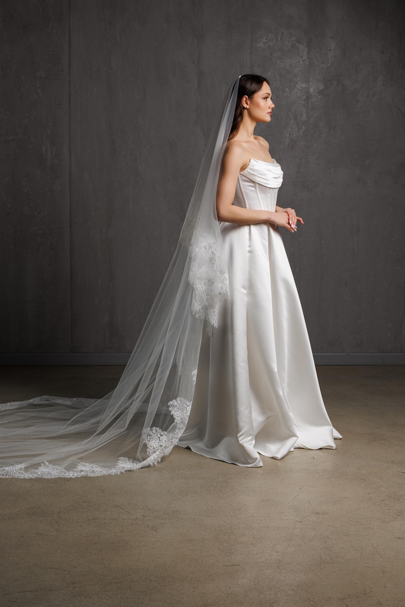 Puede incluir: Vestido de novia de sat&eacute;n blanco con corpi&ntilde;o sin tirantes y falda en l&iacute;nea A. Un velo largo y transparente con ribete de encaje cae por la espalda. El vestido es elegante y cl&aacute;sico.