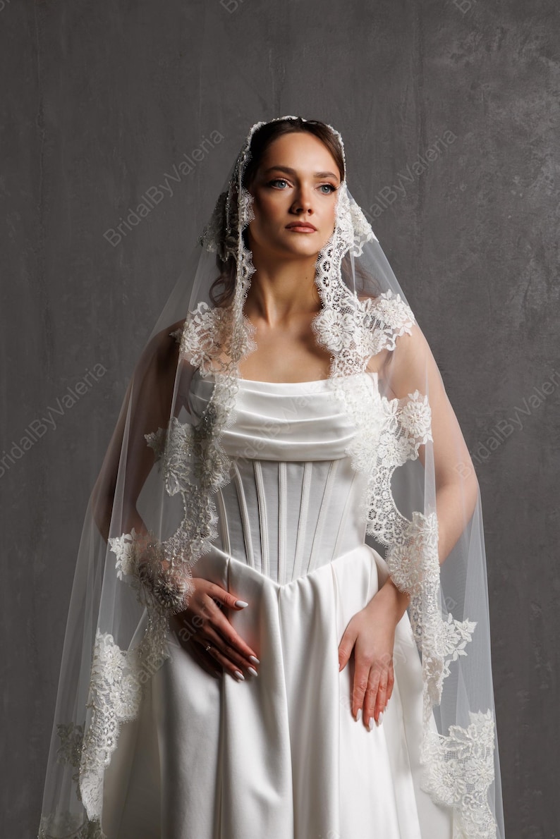 May include: A white wedding dress with a corset bodice and a flowing skirt. The dress is paired with a sheer veil trimmed with delicate lace. The image is set against a neutral gray backdrop.