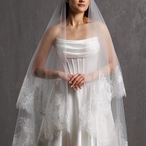 Puede incluir: Un vestido de novia blanco con un corpi&ntilde;o sin tirantes y una falda fluida. Un velo transparente de dos niveles con delicados adornos de encaje cubre los hombros. La novia est&aacute; de pie frente a un fondo gris.