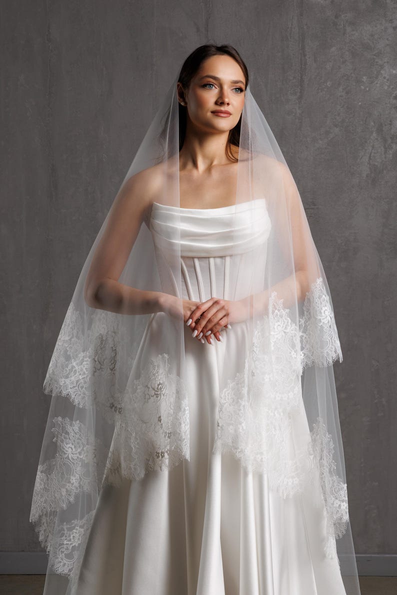 Puede incluir: Un vestido de novia blanco con un corpi&ntilde;o sin tirantes y una falda fluida. Un velo transparente de dos niveles con delicados adornos de encaje cubre los hombros. La novia est&aacute; de pie frente a un fondo gris.