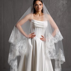 Puede incluir: Un vestido de novia blanco con un corpi&ntilde;o sin tirantes y una falda fluida. El vestido se combina con un velo transparente adornado con delicado encaje. La novia est&aacute; de pie con las manos en las caderas.