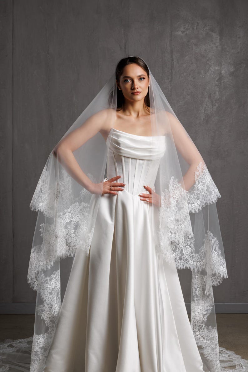 Puede incluir: Un vestido de novia blanco con un corpi&ntilde;o sin tirantes y una falda fluida. El vestido se combina con un velo transparente adornado con delicado encaje. La novia est&aacute; de pie con las manos en las caderas.