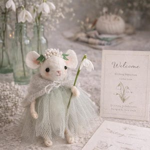 May include: A small white felted mouse figurine wearing a light gray cape and a pale green tulle skirt. The mouse has a floral crown of tiny white beads and green leaves, and holds a single white snowdrop flower. Two instructional cards are present.