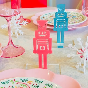 Op de afbeelding: Twee notenkraker-kaartjeshouders, een roze en een blauwe, op een wit tafelkleed met roze en witte borden. De notenkrakers zijn gemaakt van acryl en hebben een speels ontwerp.