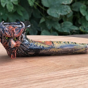 May include: A ceramic pipe shaped like a grotesque face with green eyes, a red and orange beard, and a black and green body.