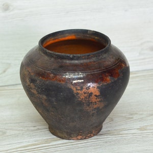May include: A dark brown clay pot with a wide mouth and a rounded body. The pot has a worn, rustic appearance with a hint of orange glaze.