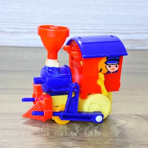 May include: A colorful toy train with a red engine, blue accents, and a blue roof. The train has yellow wheels and a smiling conductor in the window. The toy is made of plastic and sits on a wooden surface.