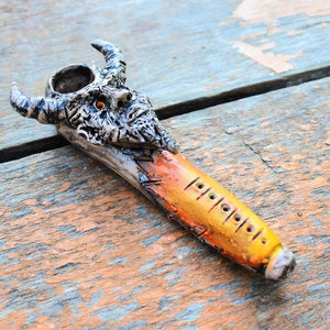 May include: A ceramic pipe shaped like a demon's head with horns. The pipe is painted in shades of gray, black, orange, and yellow. The pipe has a small bowl and a long, thin stem with holes along the side.