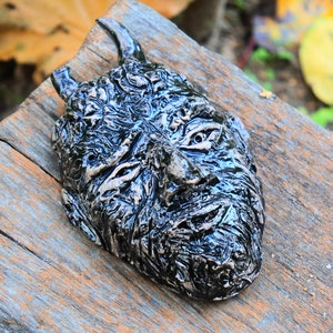 Op de afbeelding: Een zwart keramiek masker met een getextureerd oppervlak en een paar hoorns. Het masker heeft een gedetailleerd gezicht met een gefronste wenkbrauw en een licht geopende mond.