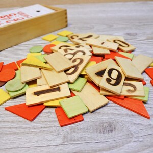 以下が含まれることがあります： カラフルな木製の形と数字のタイルが入った木製の箱。箱には「Color & Count Game」とラベルが貼られています。