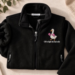 May include: A black fleece jacket with a full-length zipper and a high collar. The chest features an embroidered white duck wearing pink boxing gloves, accompanied by the text "Off to fight the bad cells" in white lettering.