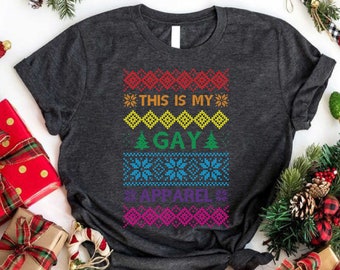 May include: A dark grey t-shirt with a rainbow-coloured Christmas jumper design. The design features the text "This is my Gay Apparel" with snowflakes and Christmas trees.