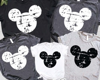 Könnte beinhalten: Fünf T-Shirts in verschiedenen Farben: Schwarz, Grau, Dunkelgrau, Weiß und Hellgrau. Jedes Shirt hat eine weiße Mickey Mouse Kopf Grafik im Used-Look. Die Shirts sind kurzärmlig und haben einen Rundhalsausschnitt.