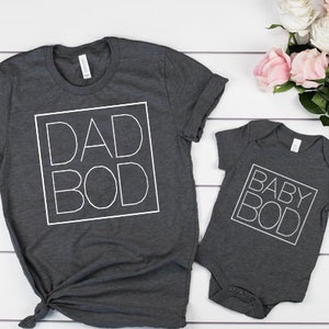 以下が含まれることがあります： 「DAD BOD」と「BABY BOD」の白いブロック文字がプリントされた、大人用とベビー用の2枚のチャコールグレーのTシャツ。ピンクと白のバラの花束が白い木の表面に置かれています。