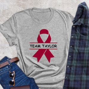 May include: A gray t-shirt with a red ribbon and the text "TEAM TAYLOR" printed on it. The shirt is laid out on a light gray surface with a pair of blue jeans, a brown belt, sunglasses, and a brown case.