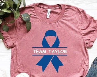 Op de afbeelding: Een heide-paarse t-shirt met een blauw lint en de tekst "TEAM TAYLOR" bedrukt op de voorkant.