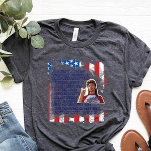 Op de afbeelding: Donkergrijs t-shirt met een grafisch ontwerp met een Amerikaanse vlag en een portret van een man. De tekst op het shirt luidt: "YOU DON'T HAVE NO WHISTLIN' BUNGHOLES..." en gaat verder met een lijst met humoristische zinnen. Het shirt wordt getoond met een spijkerbroek en sandalen.