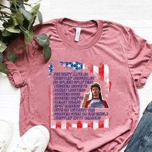 Op de afbeelding: Heather mauve t-shirt met een afbeelding met een man en tekst in een patriottisch ontwerp. De tekst luidt: "YOU DON'T HAVE NO WHISTLIN' BUNGHOLES...WHISTLIN' KITTY CHASER?" Het shirt wordt gecombineerd met een blauwe jeans en witte sneakers.