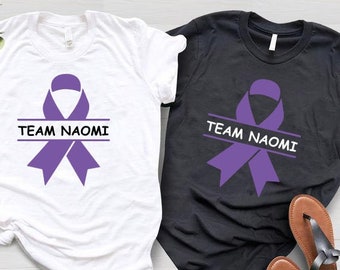Op de afbeelding: Twee T-shirts met een paars lint en de tekst "Team Naomi" erop. Een shirt is wit en de andere is zwart.