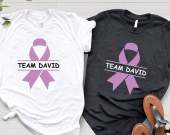 Op de afbeelding: Twee T-shirts met een paars lint en de tekst "Team David" erop. Een shirt is wit en de andere is zwart.