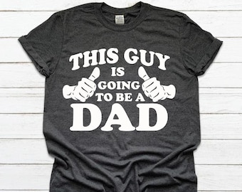May include: Dark gray t-shirt with white text and thumbs-up graphic. The text reads "THIS GUY IS GOING TO BE A DAD". A fun and celebratory design for expecting fathers.