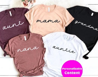 May include: Five different t-shirts with various family member titles written in cursive. The shirts are in different colors, including maroon, peach, black, brown, and white. The text on the shirts reads "aunt", "mama", "grandma", "nana", and "auntie".