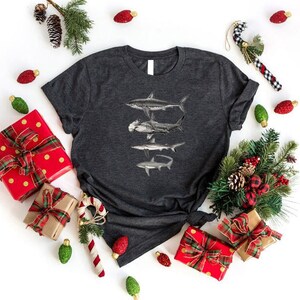 May include: A dark gray t-shirt with a graphic of three sharks in a stacked formation. The sharks are black and white and are silhouetted against a gray background. The shirt is surrounded by Christmas decorations, including red and gold gift boxes, green pine branches, and red and white candy canes.
