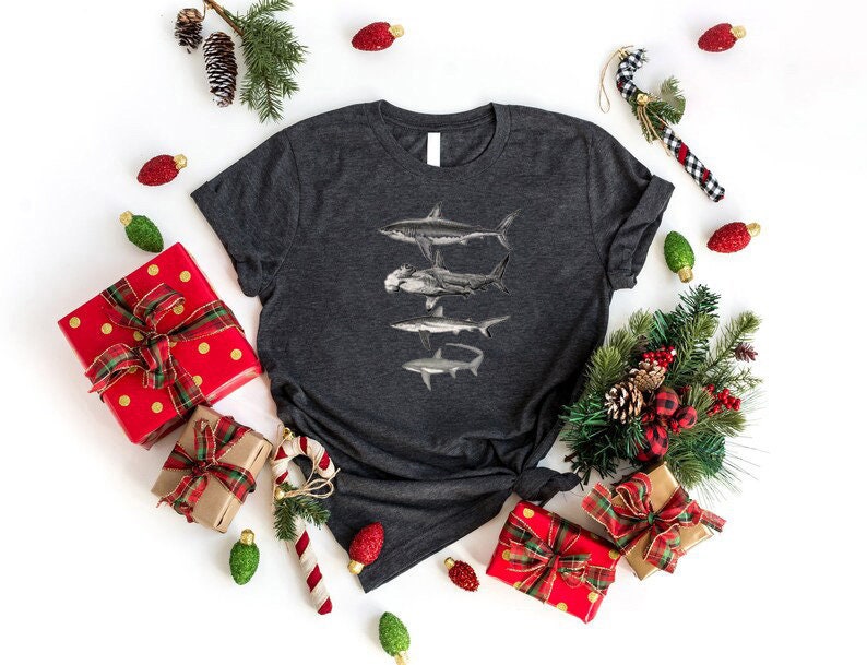 May include: A dark gray t-shirt with a graphic of three sharks in a stacked formation. The sharks are black and white and are silhouetted against a gray background. The shirt is surrounded by Christmas decorations, including red and gold gift boxes, green pine branches, and red and white candy canes.