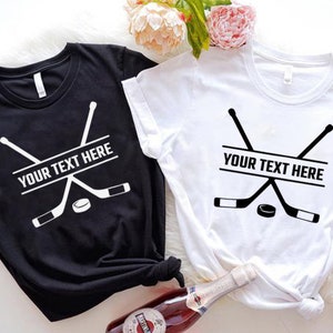 Może przedstawiać: Dwie koszulki z nadrukiem hokejowej kija i krążka. Na obu koszulkach widnieje napis "YOUR TEXT HERE". Jedna koszulka jest czarna, a druga biała.
