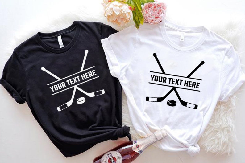 Może przedstawiać: Dwie koszulki z nadrukiem hokejowej kija i krążka. Na obu koszulkach widnieje napis "YOUR TEXT HERE". Jedna koszulka jest czarna, a druga biała.