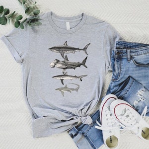 May include: A gray t-shirt with a graphic of four different types of sharks. The sharks are drawn in black and white and are arranged vertically on the shirt. The shirt is folded and laid on a white surface with a pair of blue jeans and white sneakers.