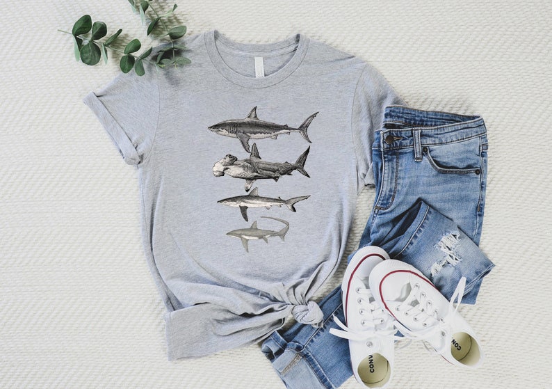 May include: A gray t-shirt with a graphic of four different types of sharks. The sharks are drawn in black and white and are arranged vertically on the shirt. The shirt is folded and laid on a white surface with a pair of blue jeans and white sneakers.