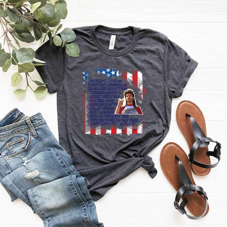 Op de afbeelding: Donkergrijs t-shirt met een grafisch ontwerp met een Amerikaanse vlag en een portret van een man. De tekst op het shirt luidt: "YOU DON'T HAVE NO WHISTLIN' BUNGHOLES..." en gaat verder met een lijst met humoristische zinnen. Het shirt wordt getoond met een spijkerbroek en sandalen.
