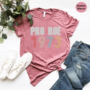 May include: A heather mauve t-shirt with the text "PRO ROE 1973" in a retro font. The numbers are in different colors, with red, orange, blue, and yellow.