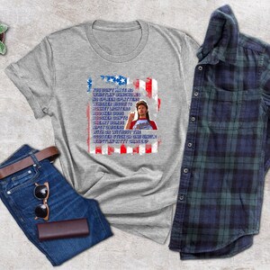 Op de afbeelding: Een grijs t-shirt met een afbeelding van een Amerikaanse vlag en tekst. De afbeelding bevat een spijkerbroek, een bruine riem, een zonnebril, een portemonnee en een geruit overhemd. Een kleine potplant is ook zichtbaar.